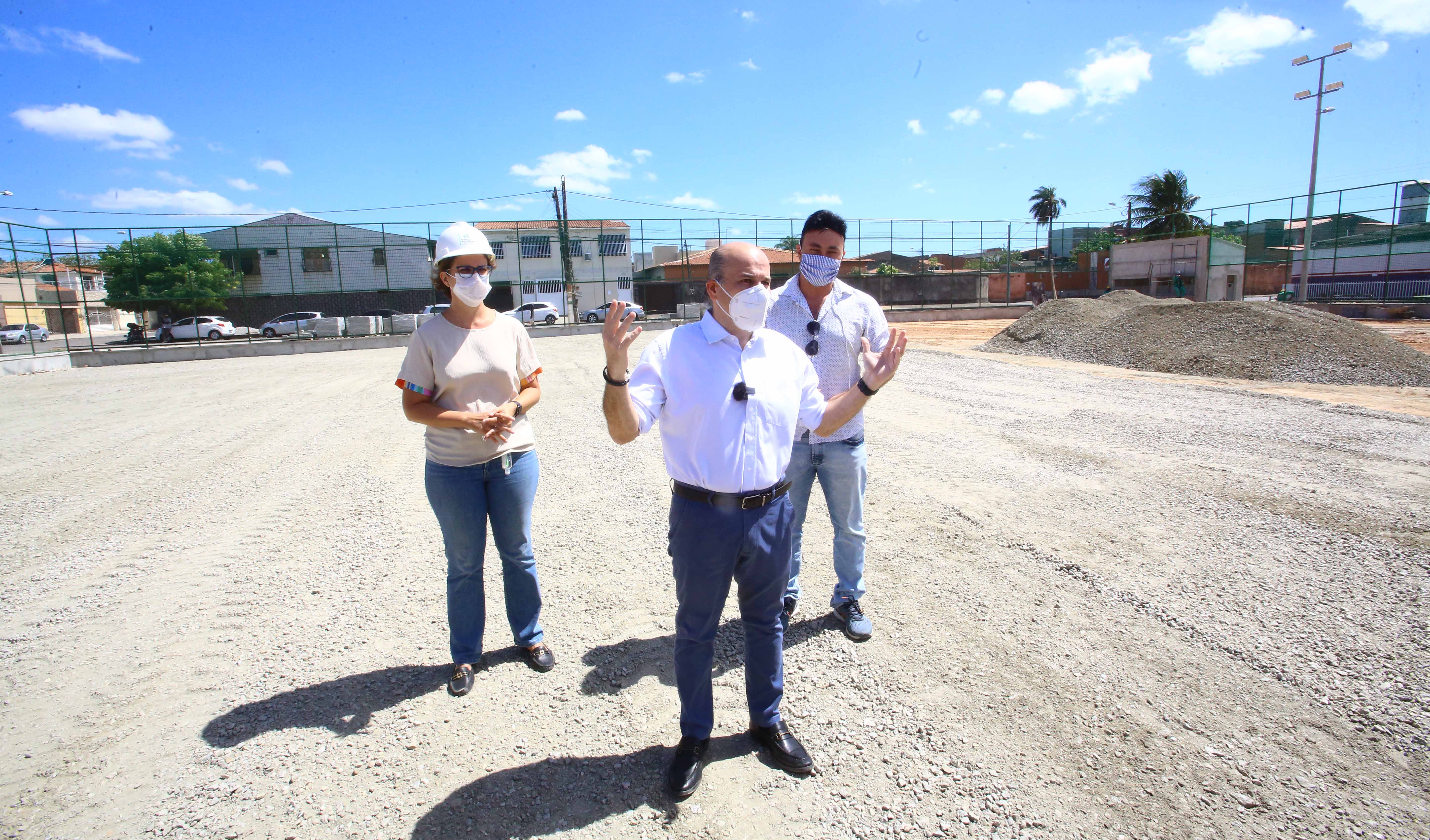 prefeito em pé ao lado de secretária e homem, todos de máscara com obra em campo de areia em obras ao fundo